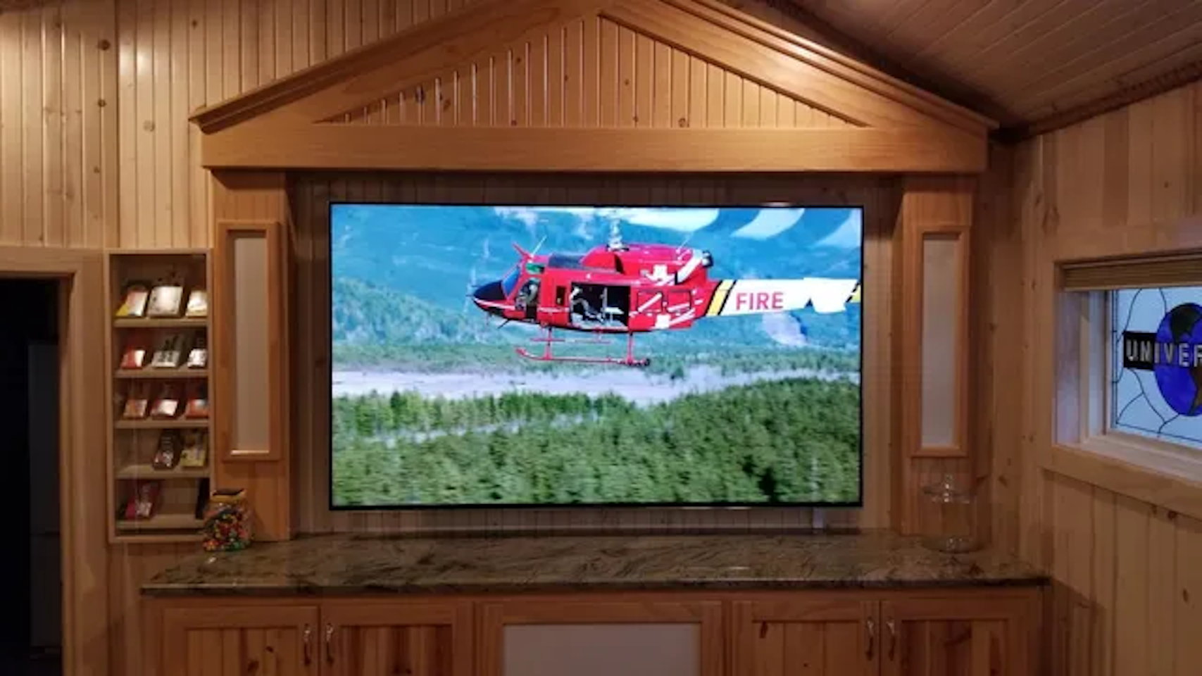 A home theater TV screen showing a red helicopter flying over a forest, mounted in a wooden entertainment center.