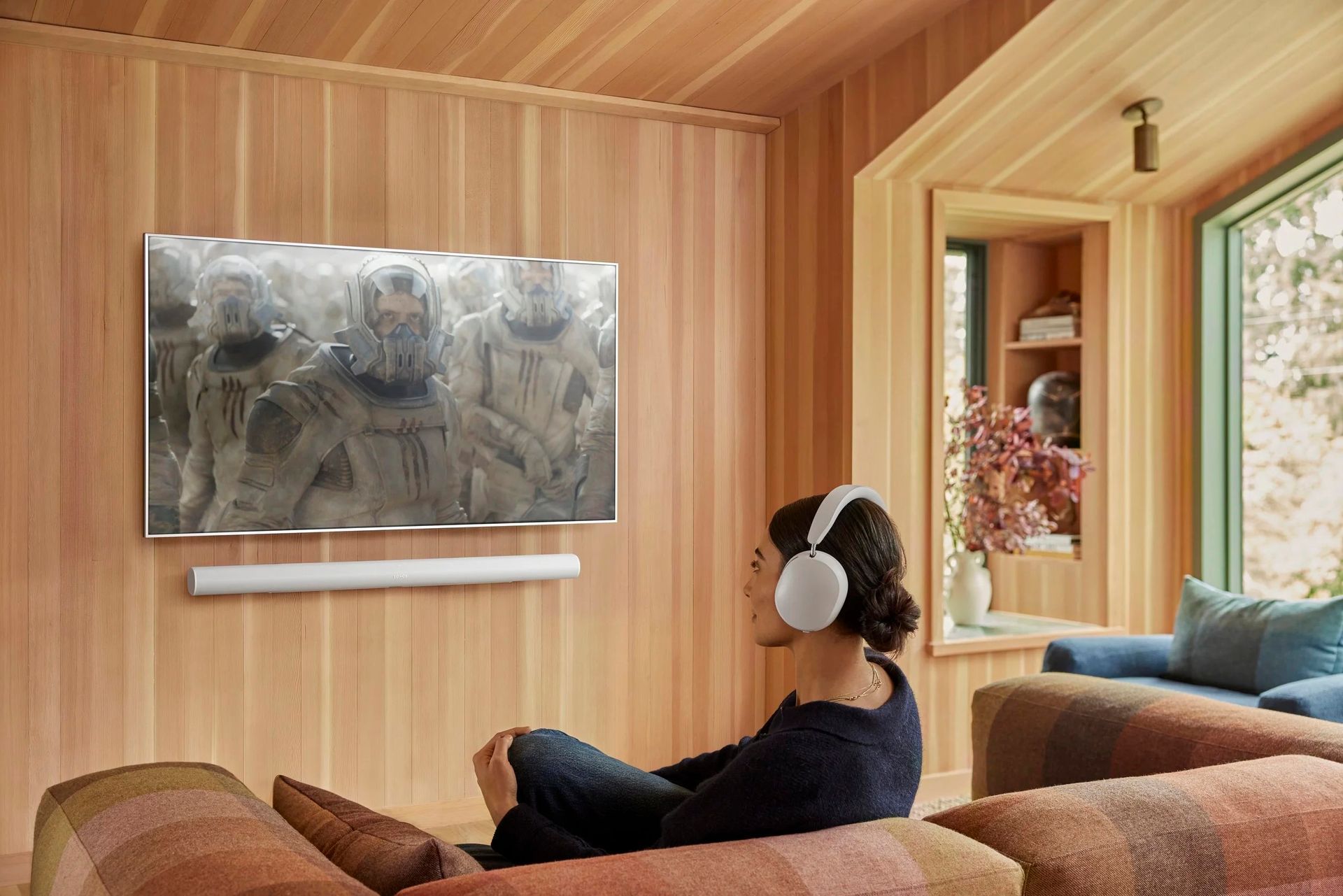 Woman wearing sonos ace headphones watches TV with a sonos arc ultra mounted on the wall.