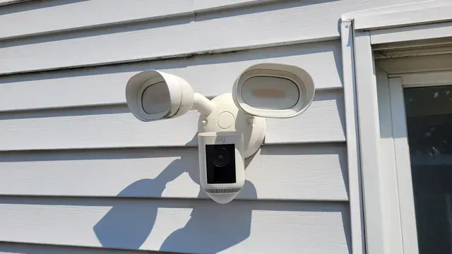 White floodlight security camera mounted on white siding next to a window.
