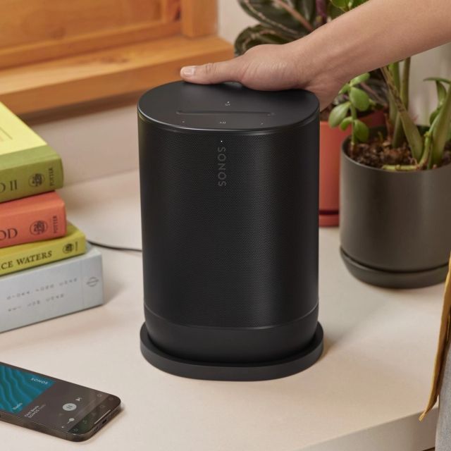 Black Sonos move 2 speaker with a hand on top, next to books and a smartphone.