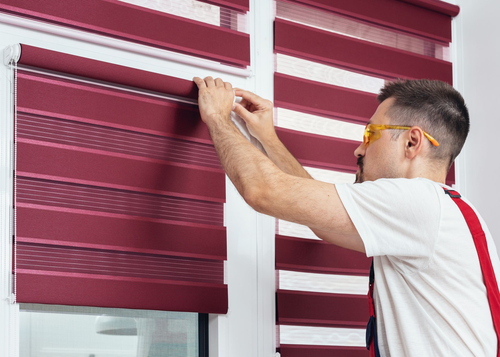 A young man in a uniform carefully installing new window blinds with focused attention.