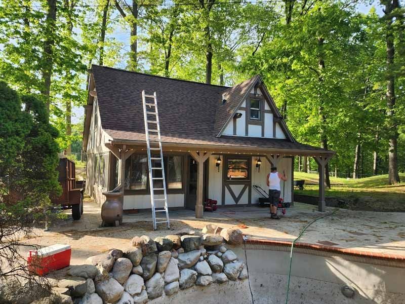 A man is standing in front of a small house with a ladder on the roof.