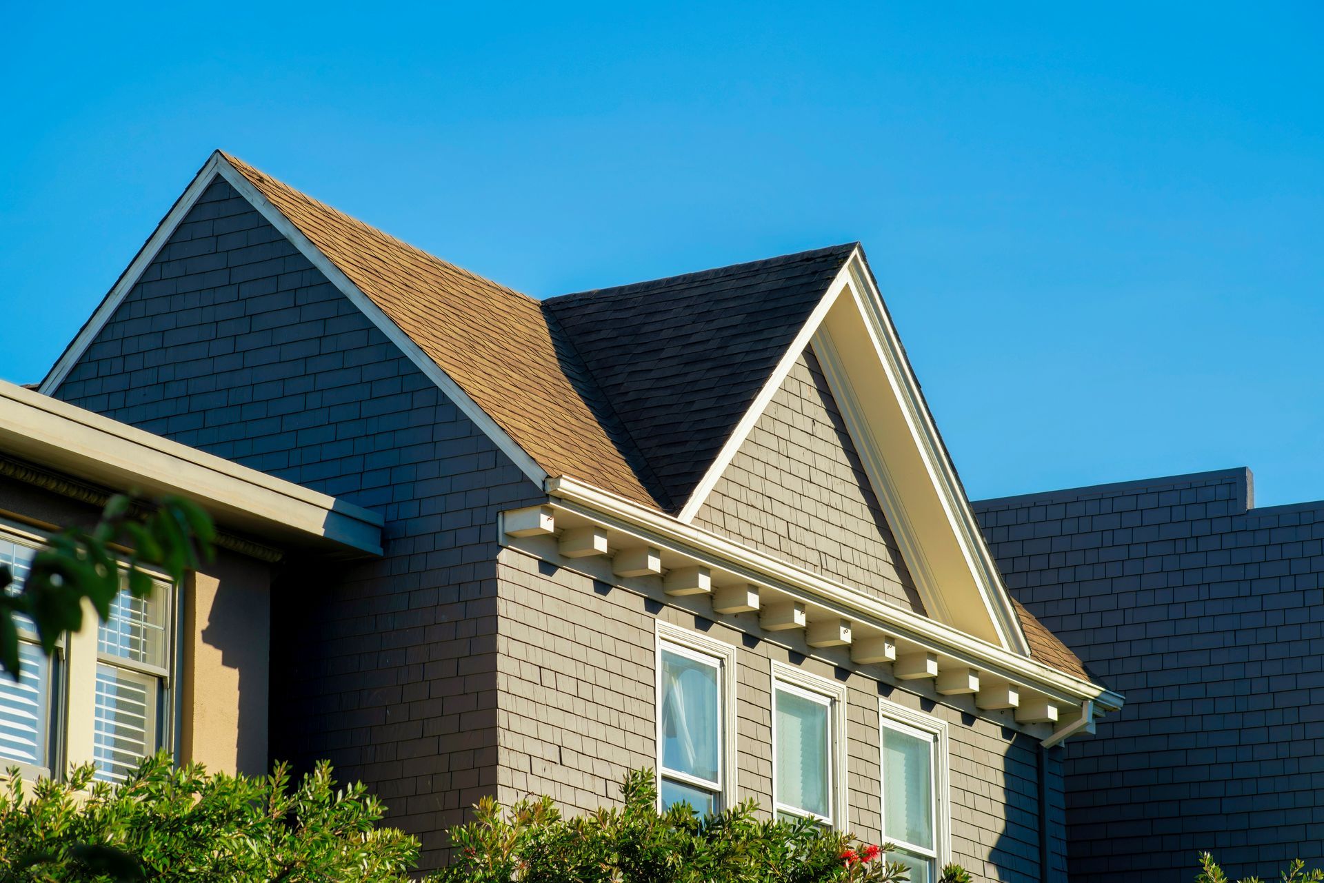 Two-story house roof with tan shingles and white trim against a blue sky.