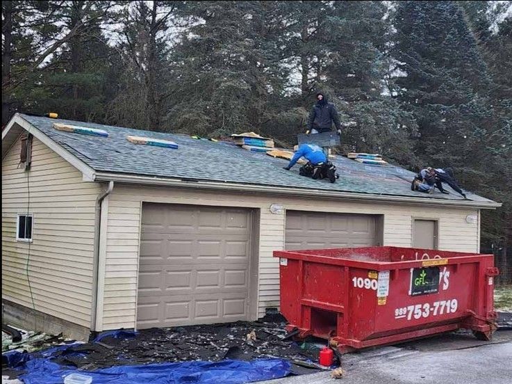 Three workers installing or repairing a shingled roof on a beige garage with two large garage doors.