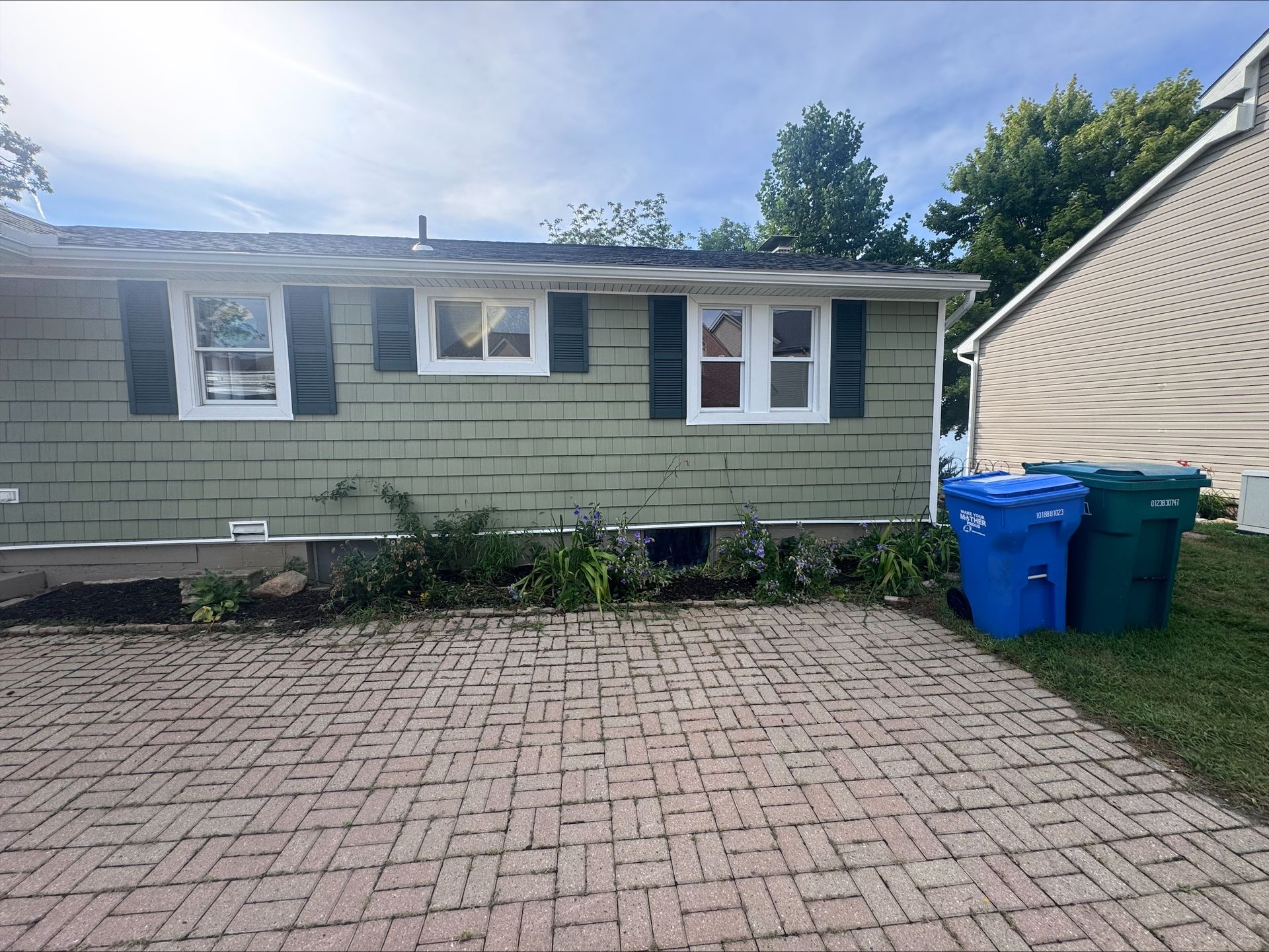 Back of a light green house with three windows, green shutters, and a brick patio. Blue and green trash bins sit on the grass.