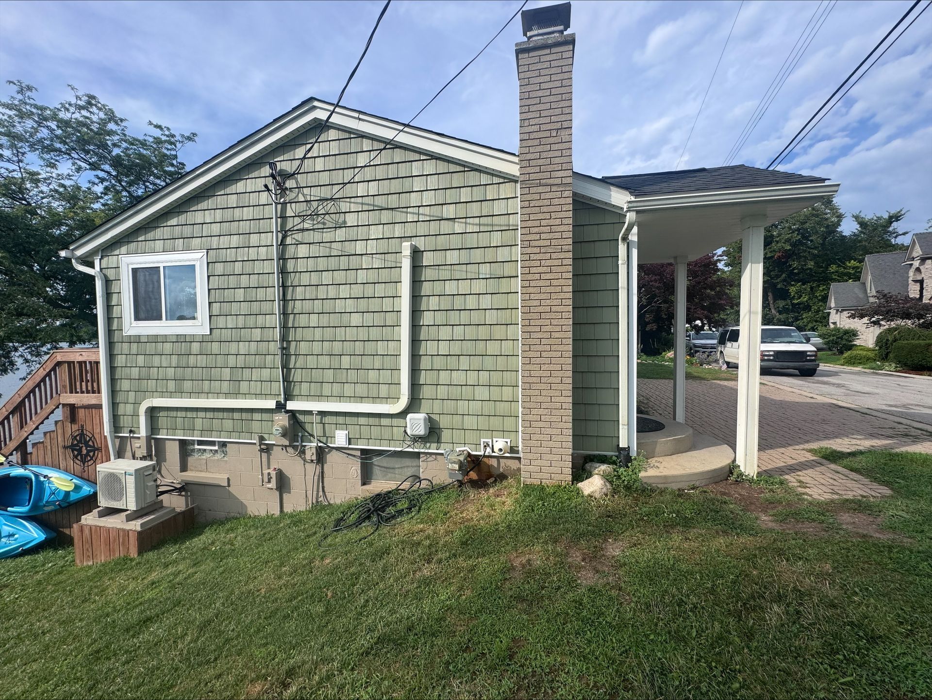 A small green house with a chimney and a covered porch on a grassy area, with a view of a street and car.