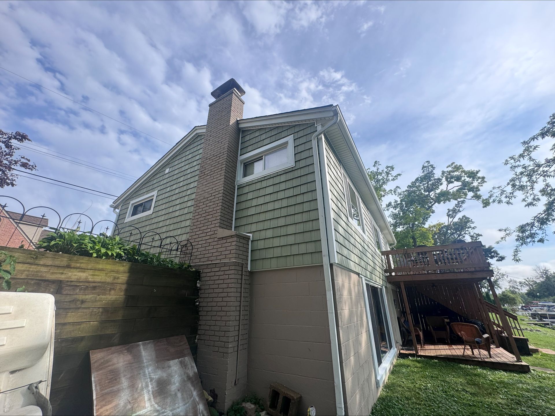 Green-sided house with a brick chimney against a cloudy sky. A wooden deck is visible on the right side.