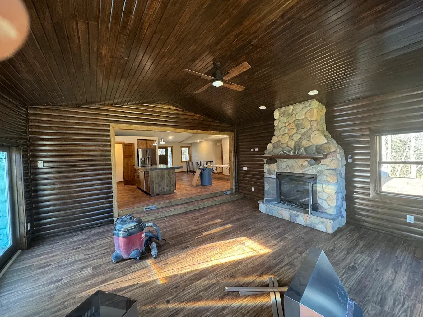 Rustic log cabin interior with stone fireplace, wood-paneled walls, and a kitchen visible through an opening