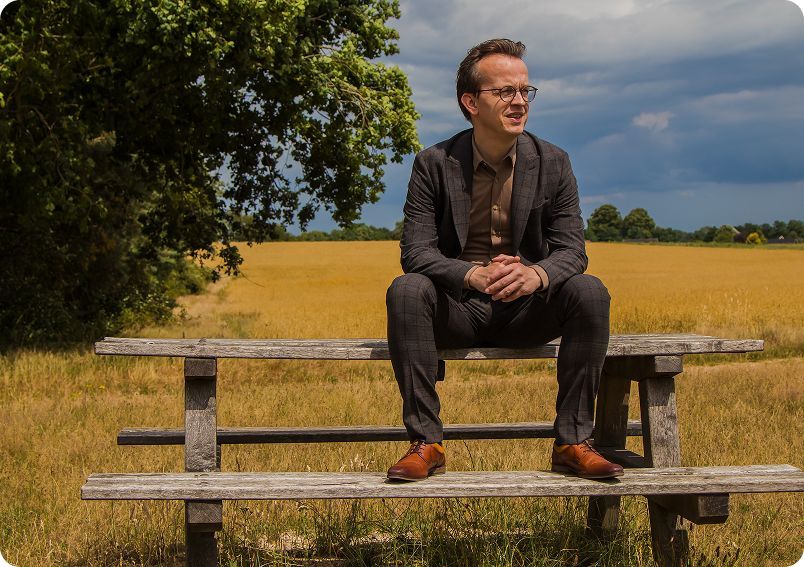 Man in suit sits on a wooden bench, looking to the side. Field and cloudy sky in background.