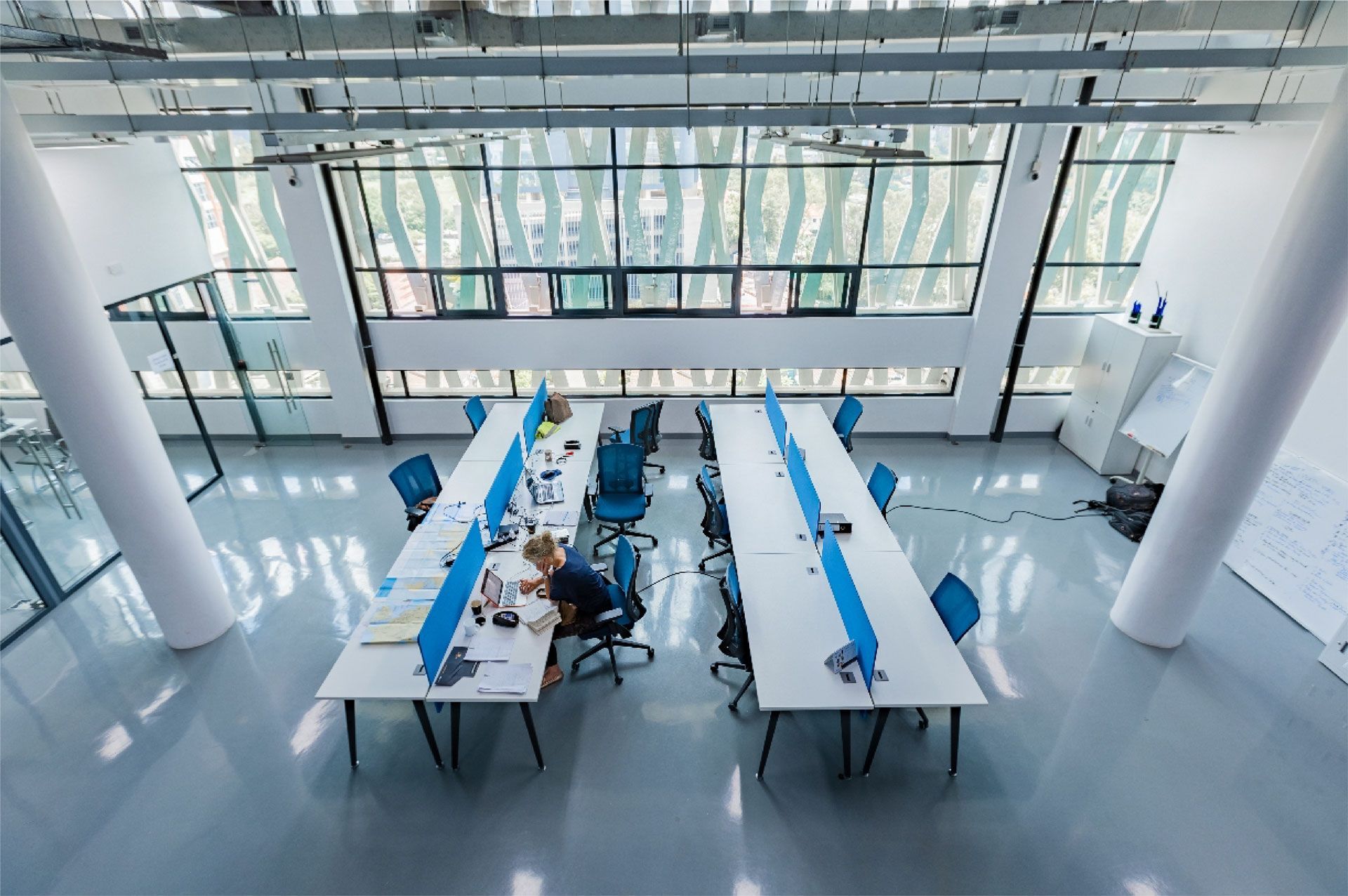 Office space with desks, blue chairs, and large windows. Someone works at a desk.