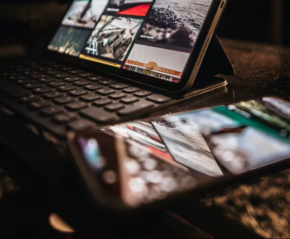 Tablet displaying images, atop a keyboard; smartphone reflects both devices on a dark surface.