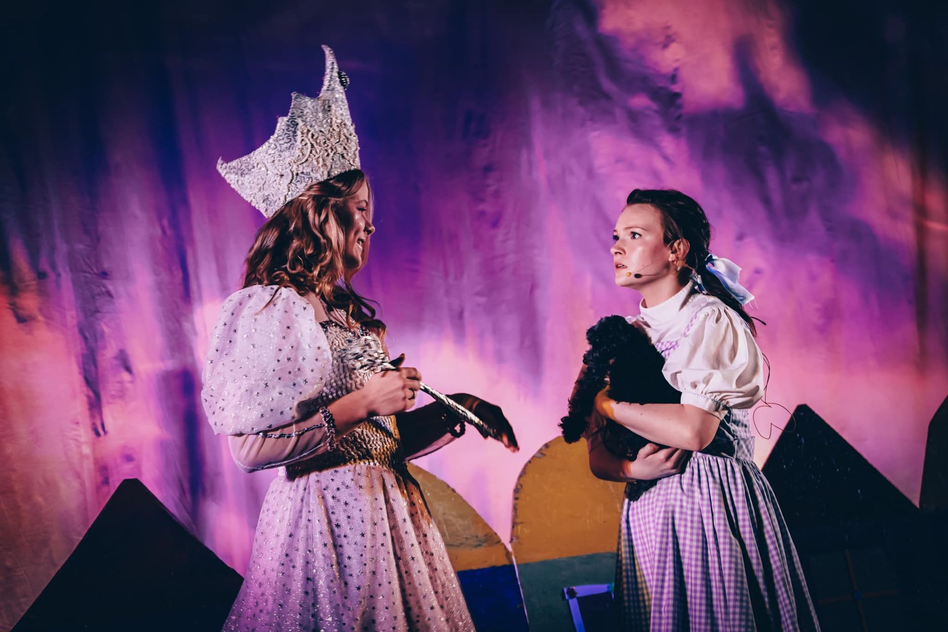 Two women on stage in costumes: one a queen with a crown, the other Dorothy from Wizard of Oz with a dog.