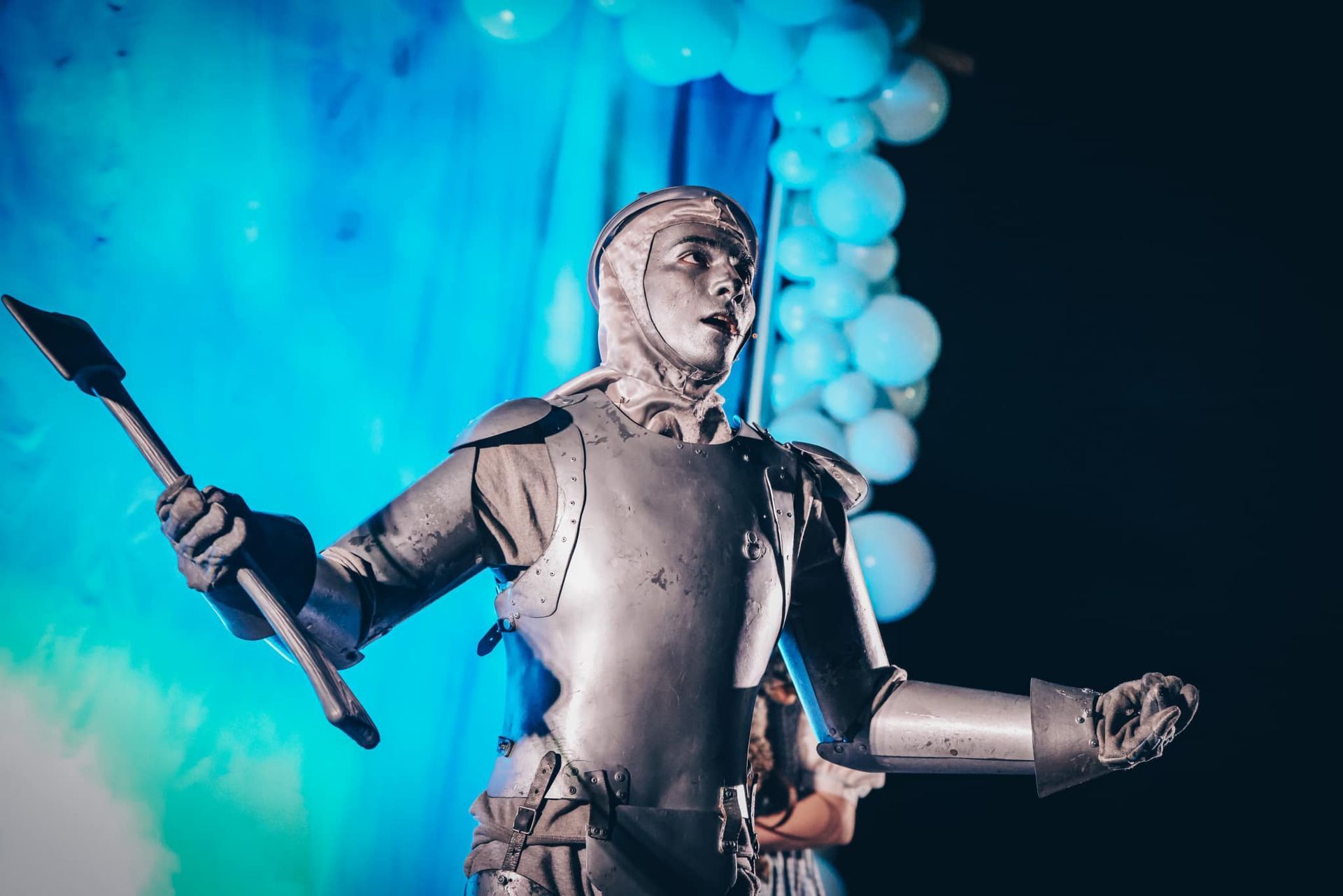 A person in a silver knight costume holds a spear, dramatic lighting, blue background and balloons.
