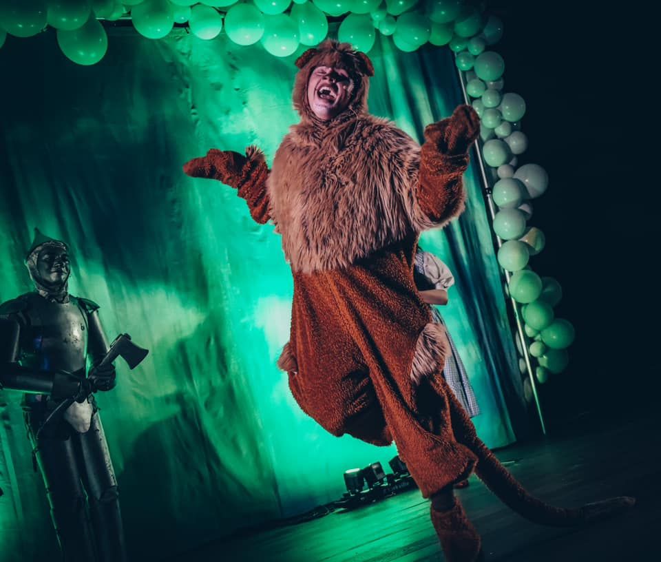 Man in Cowardly Lion costume, arms outstretched, on stage. Green backdrop, Tin Man on left.