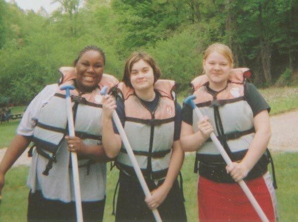 Three young women in life vests holding paddles, smiling, outdoors.