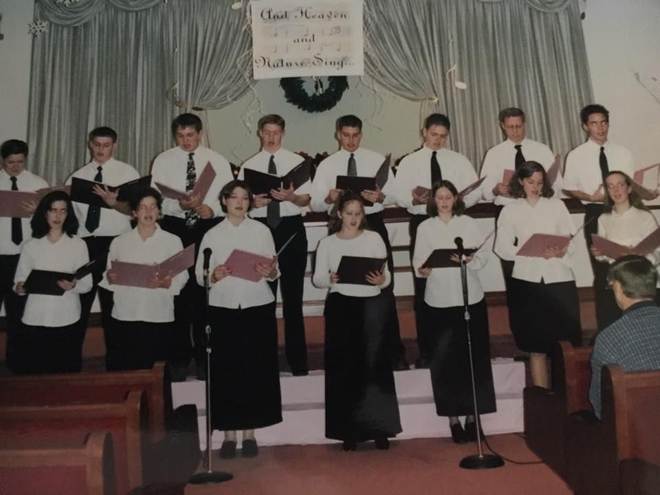Choir singing on a stage. People wearing black skirts/pants, white shirts, and ties, with a church interior as the backdrop.