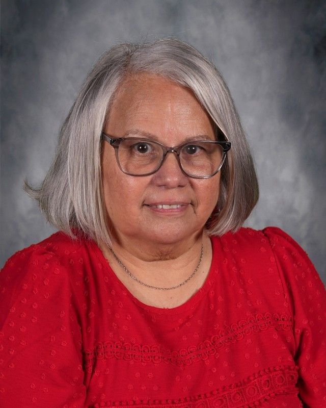 Woman with gray hair smiles, wearing a patterned blue blouse, against a blurred gray background.