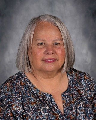 Woman with gray hair smiles, wearing a patterned blue blouse, against a blurred gray background.