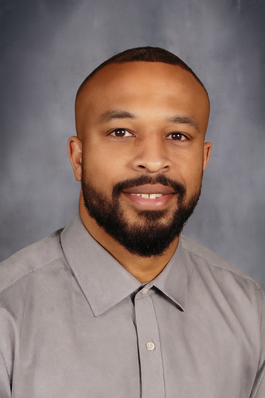 A Black man with a trimmed beard and a gray shirt smiles at the camera.