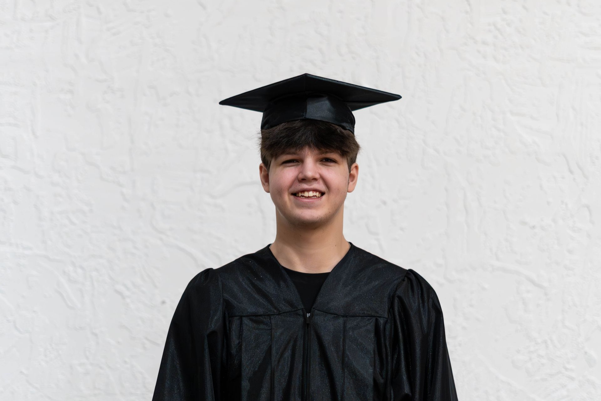 Asian graduate in cap and gown, smiling, surrounded by people, outdoor setting.