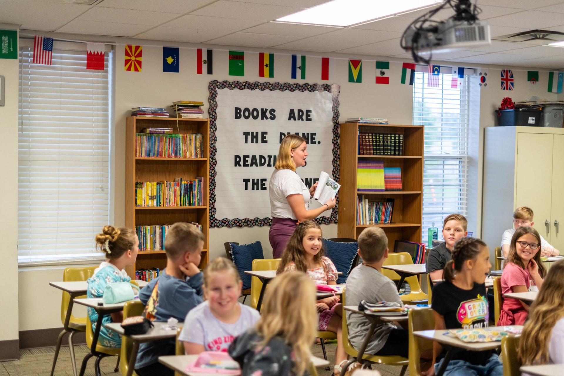 Grandmother helping granddaughter read a book at a desk; sunny interior.