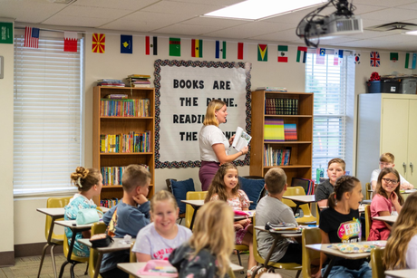 Grandmother helping granddaughter read a book at a desk; sunny interior.