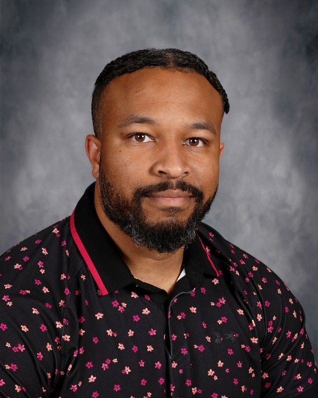 Man with brown hair and beard, smiling, wearing a pink sweater over a white patterned shirt.