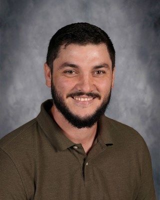 Smiling man with a full beard, wearing a brown polo shirt against a mottled gray backdrop.