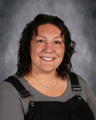 Woman with dark curly hair smiling, wearing a gray shirt and black overalls.
