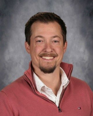 Man with brown hair and beard, smiling, wearing a pink sweater over a white patterned shirt.