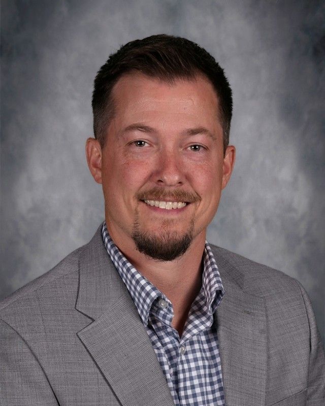 Man with brown hair and beard, smiling, wearing a pink sweater over a white patterned shirt.