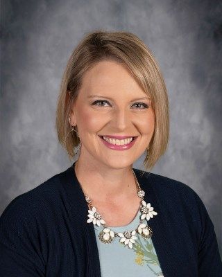 Blonde woman smiles, wearing a necklace and a blue cardigan over a light blue shirt.