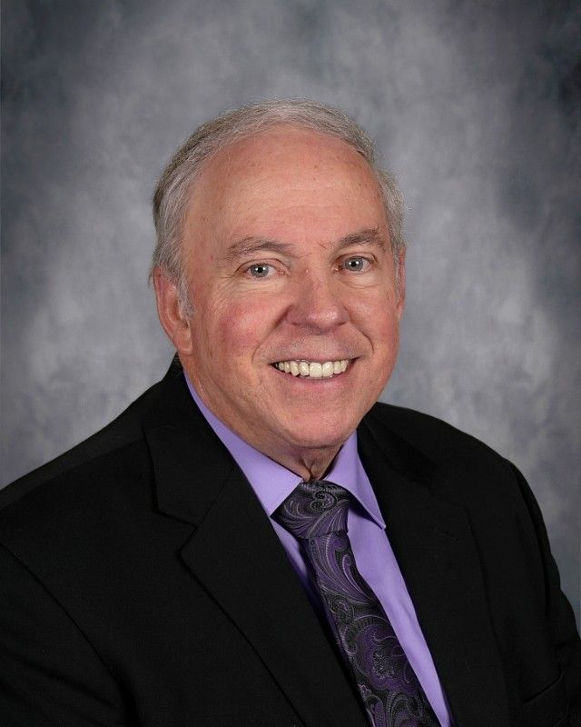 Smiling older man in a black suit and blue shirt, patterned tie, with a neutral background.