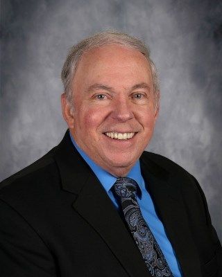 Smiling older man in a black suit and blue shirt, patterned tie, with a neutral background.