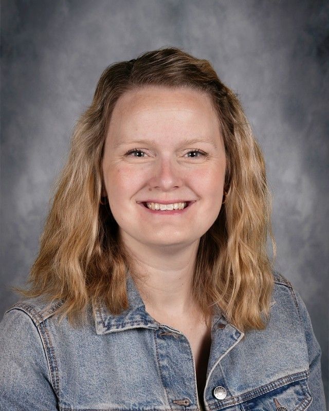 Woman with long wavy brown hair smiling, wearing a denim jacket.