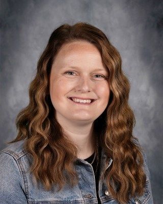 Woman with long wavy brown hair smiling, wearing a denim jacket.