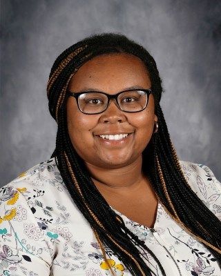 Woman with glasses and braids smiles, wearing a floral blouse, against a blurred backdrop.