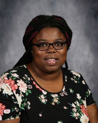 Woman with glasses and braided hair, wearing a floral top, smiles.
