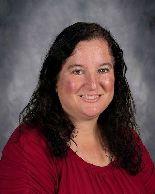 Woman with dark wavy hair, smiling, wearing a red shirt, in front of a grey backdrop.