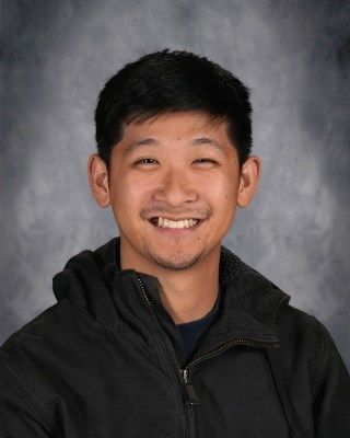 Smiling Asian man in a black hooded jacket, against a blurred gray background.