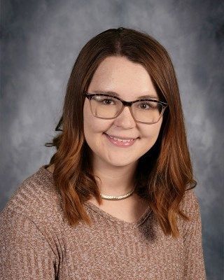 Woman with glasses smiles, wearing a brown sweater, against a gray background.