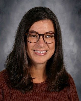 Woman with glasses, brown hair, smiling, wearing a brown sweater.