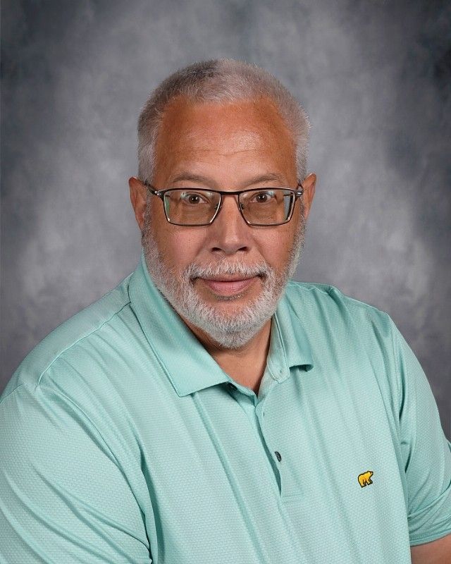 Mature man with glasses, gray beard, teal shirt smiles, on a mottled gray background.