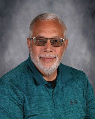 Mature man with glasses, gray beard, teal shirt smiles, on a mottled gray background.