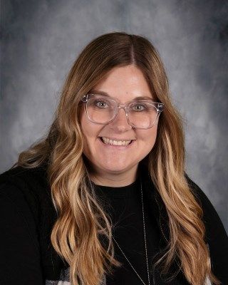 Woman with long blonde hair, glasses, and black shirt smiling, against a gray background.