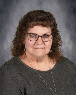 Woman with glasses, brown curly hair, and gray shirt, against a blurred gray backdrop.
