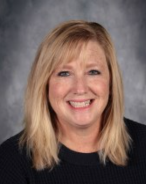 Woman with blonde hair smiling, wearing a black shirt, against a blurry gray background.