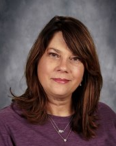 Woman with shoulder-length brown hair, wearing a purple shirt and silver necklaces; looking at the viewer.