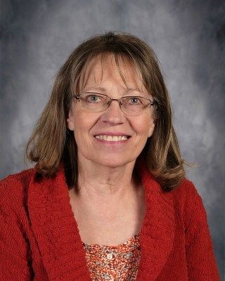 Woman with glasses, smiling, in a red sweater, against a blurred background.