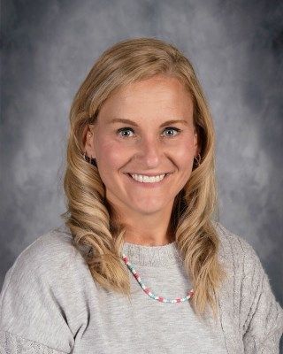 Blond woman with a bright smile, wearing a gray sweater and colorful beaded necklace, posing for a photo.